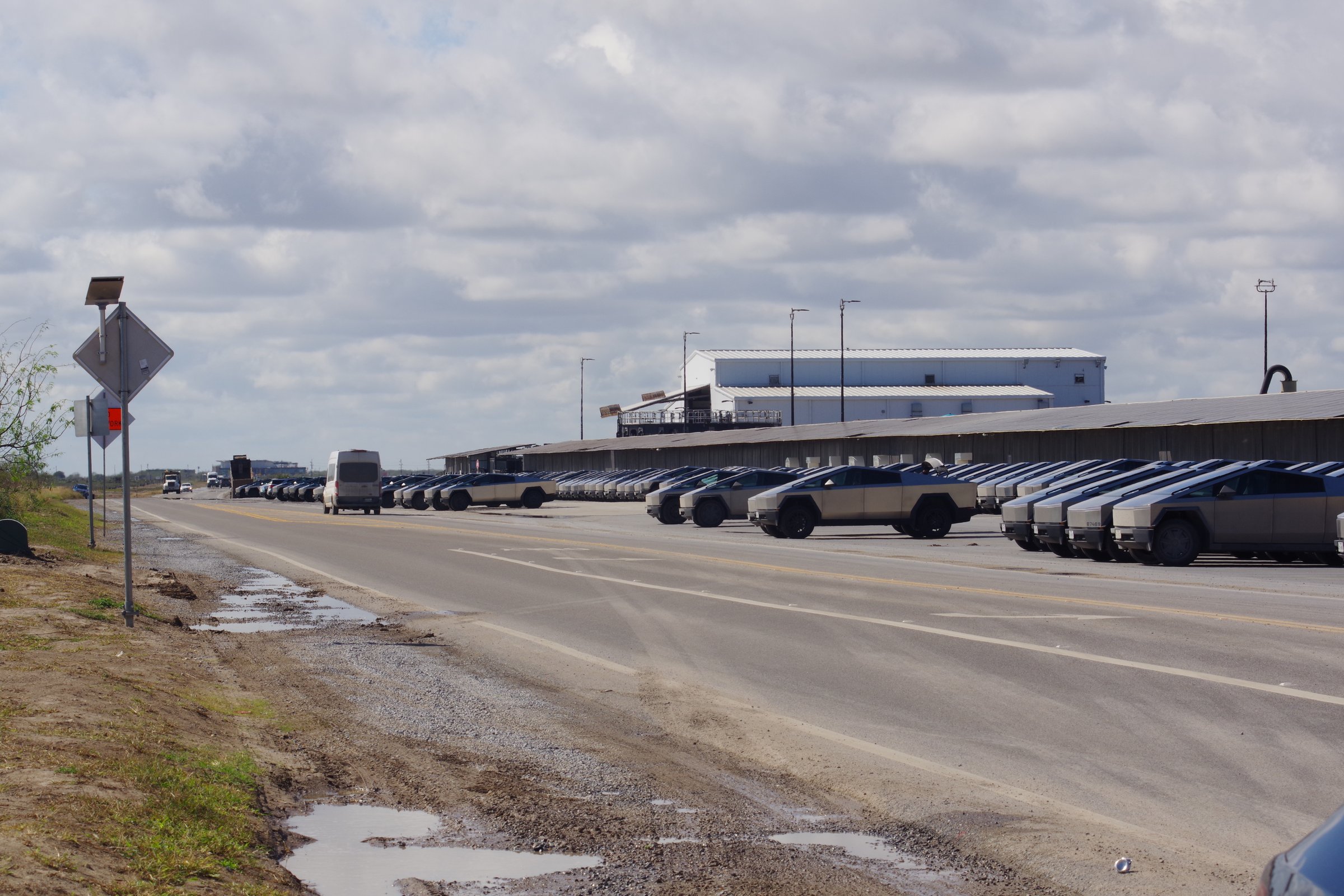 Rows of Cybertrucks in a staging lot beside a road with puddles under overcast sky