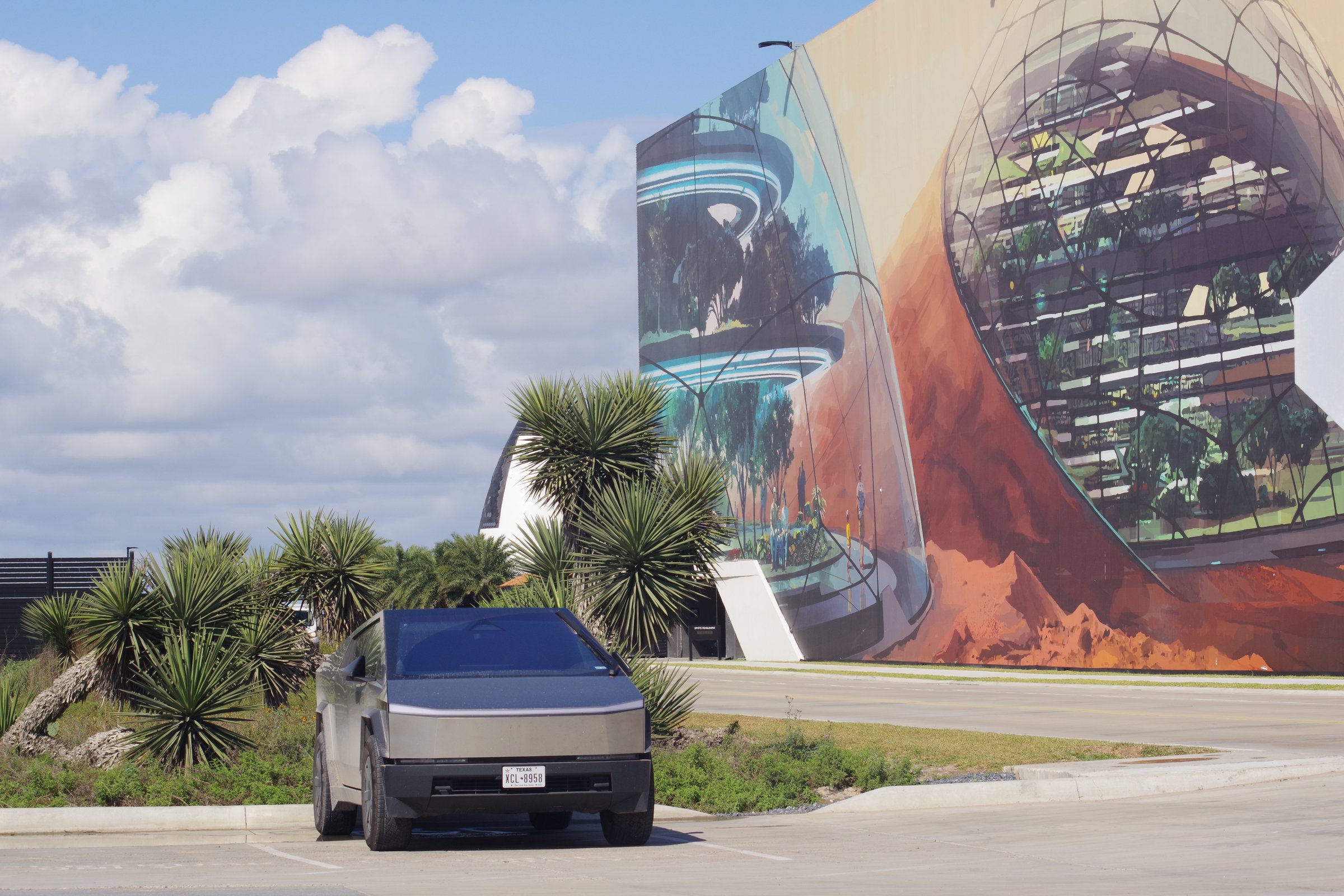 Silver Cybertruck parked before a futuristic Mars-colony mural under blue sky with yuccas