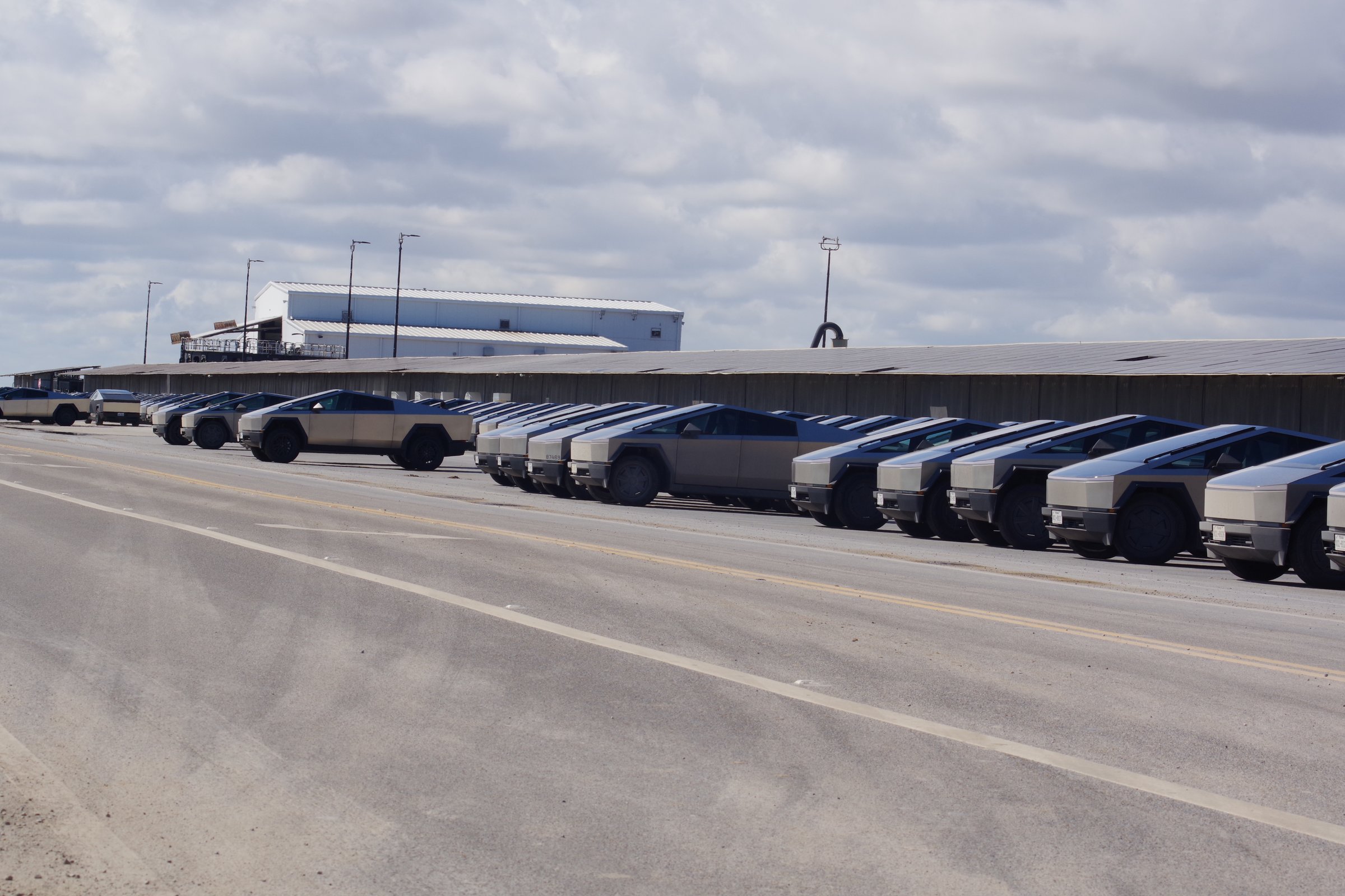 Diagonal rows of Cybertrucks under cloudy sky with an industrial building behind