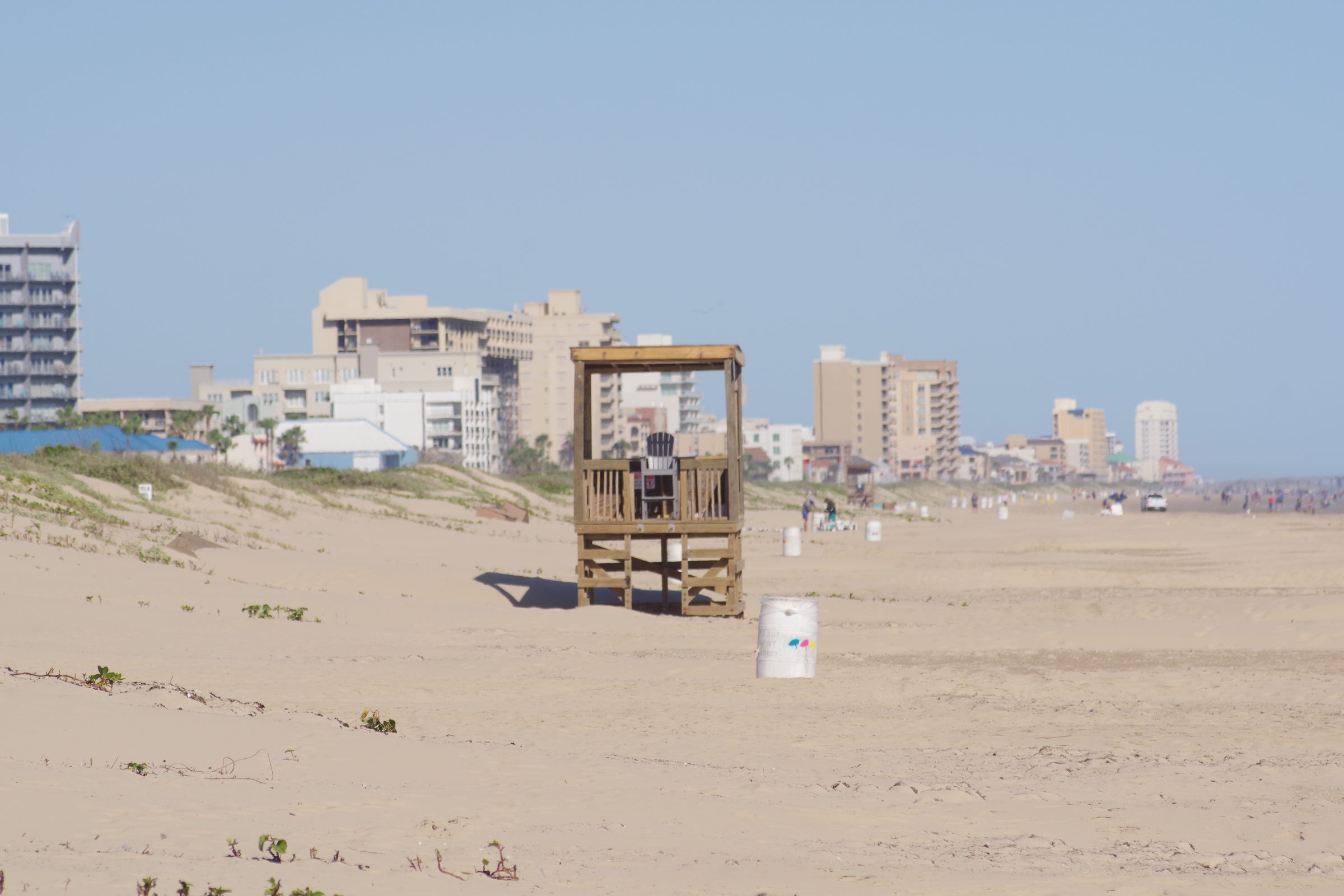 Wooden lifeguard tower on sand dunes with coastal hotels behind under pale blue sky