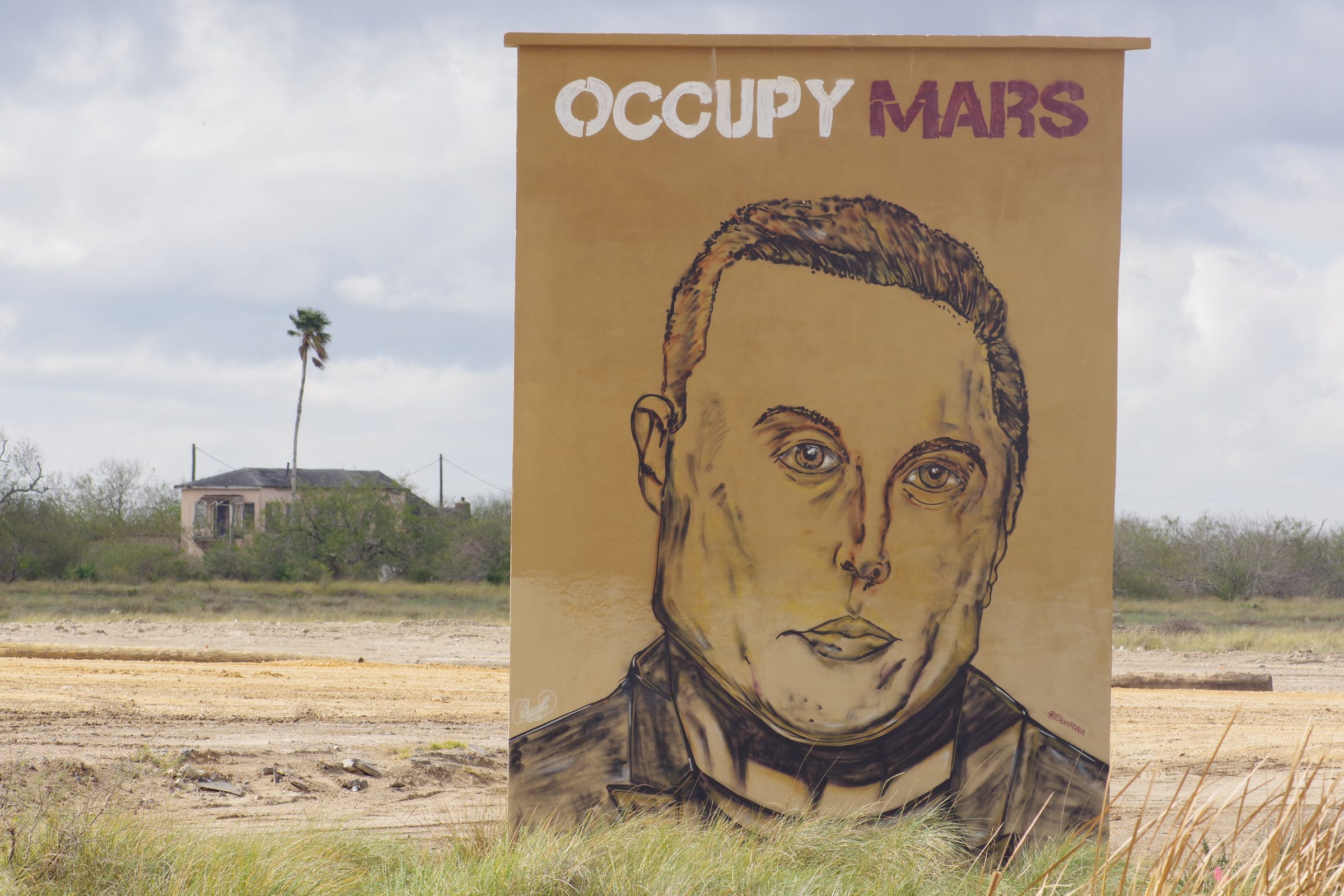 Wider view of OCCUPY MARS mural in a field with a house and palm tree in the distance