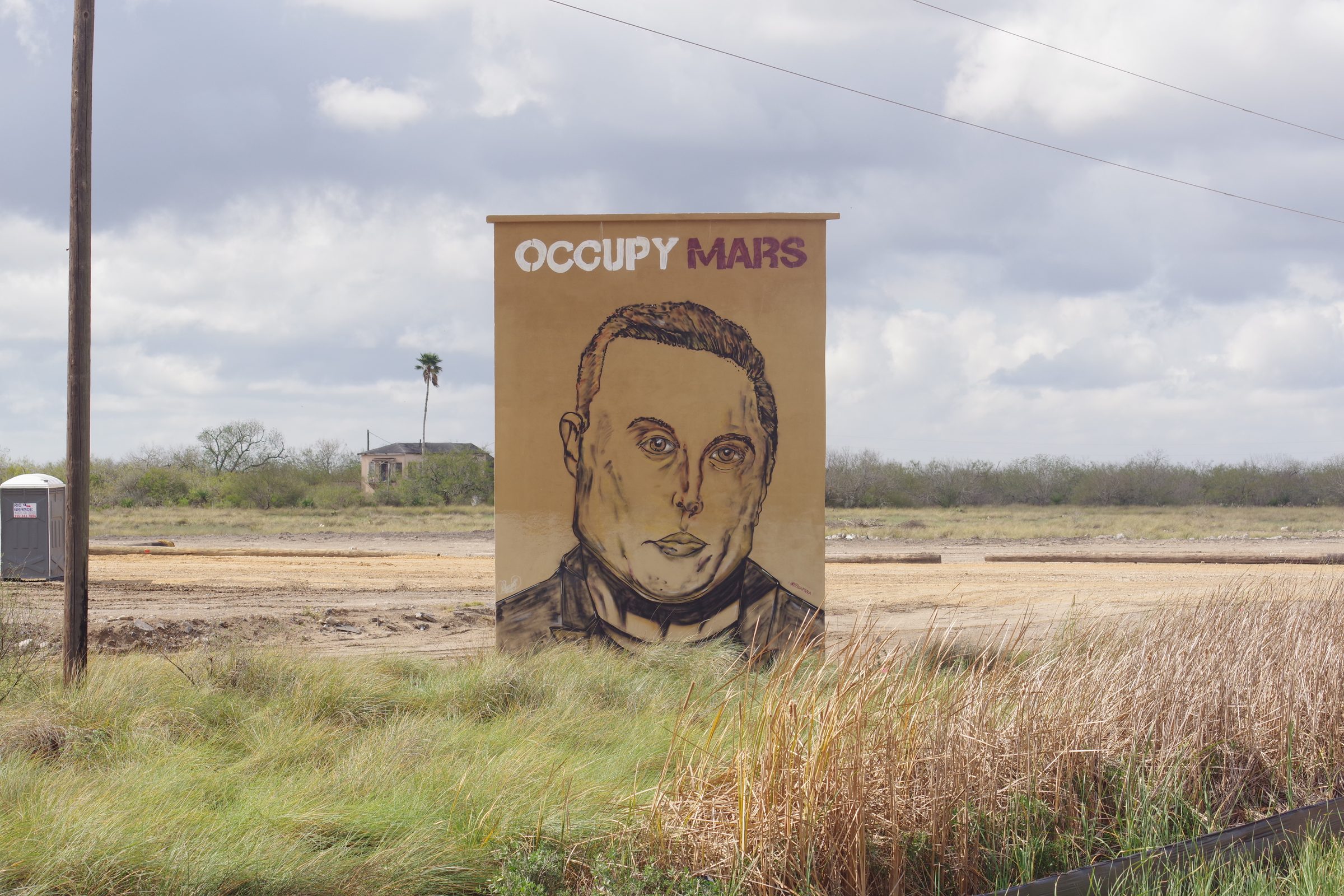 OCCUPY MARS mural with stylized Elon portrait in a field, utility pole and portable toilet visible at left
