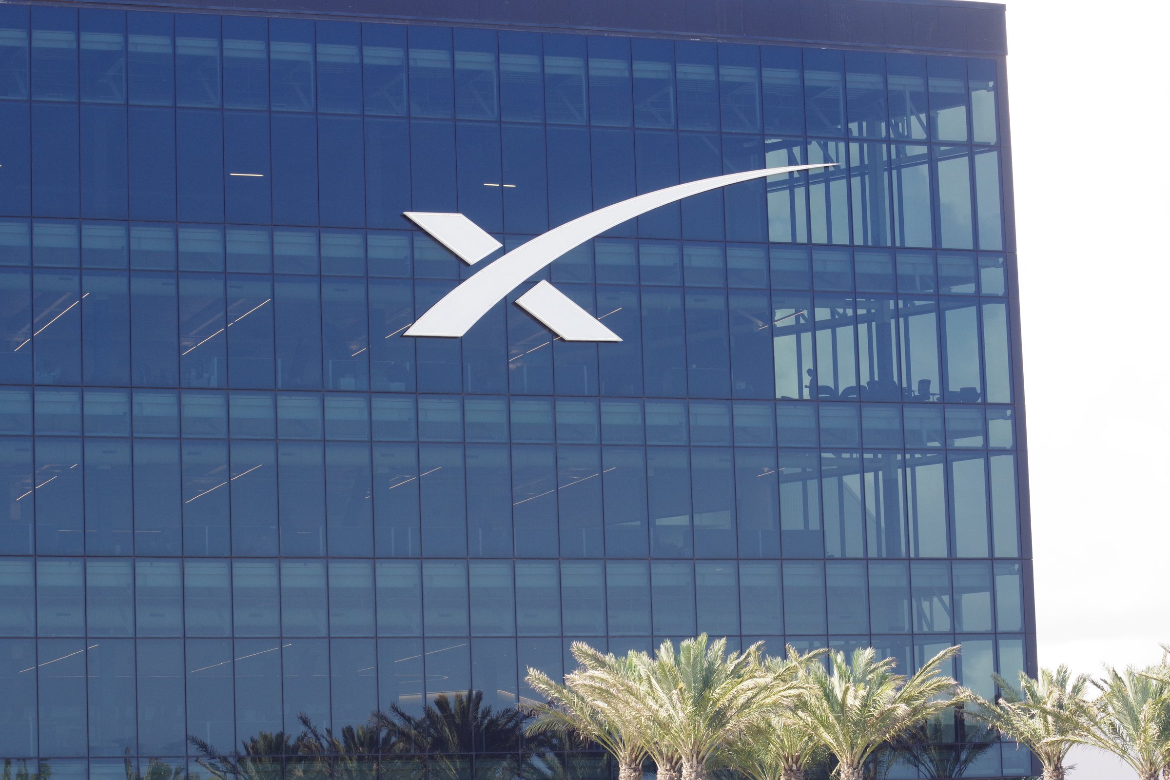 SpaceX headquarters glass facade with large white X logo and palm trees below