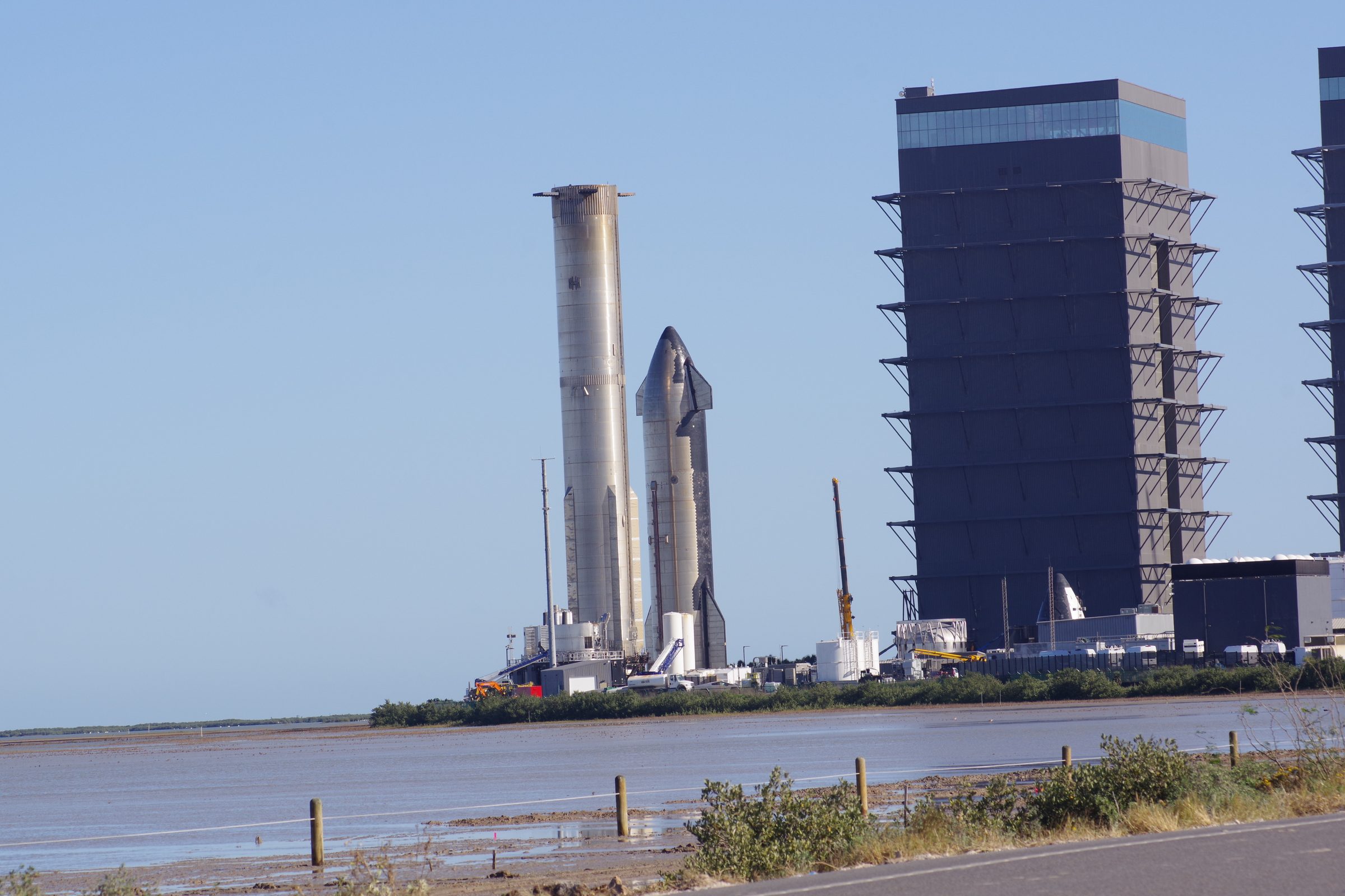 Super Heavy and Starship beside a High Bay across water, yellow crane and fence in the foreground under clear blue sky