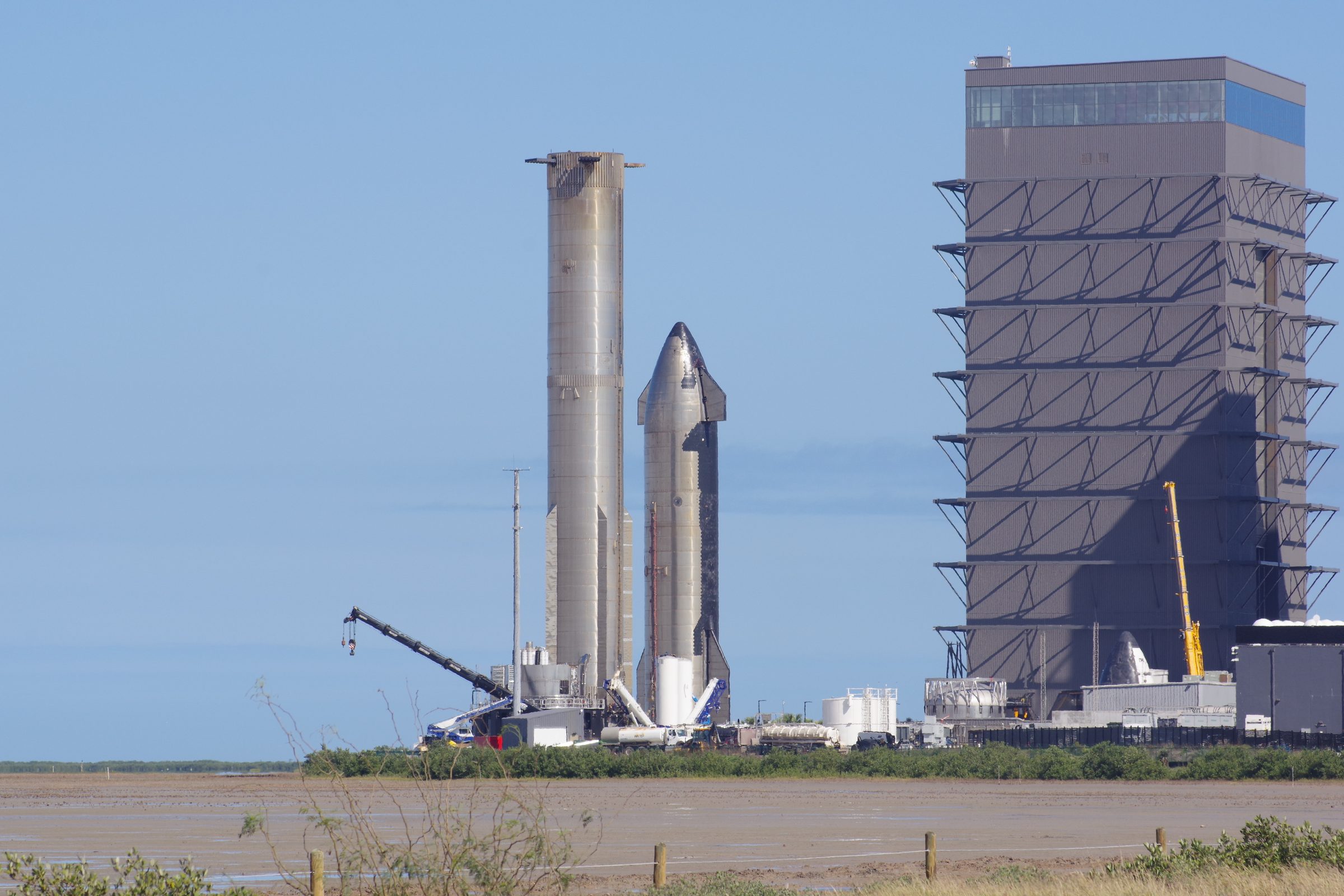 Super Heavy booster and Starship beside a High Bay under blue sky with tidal flats in the foreground