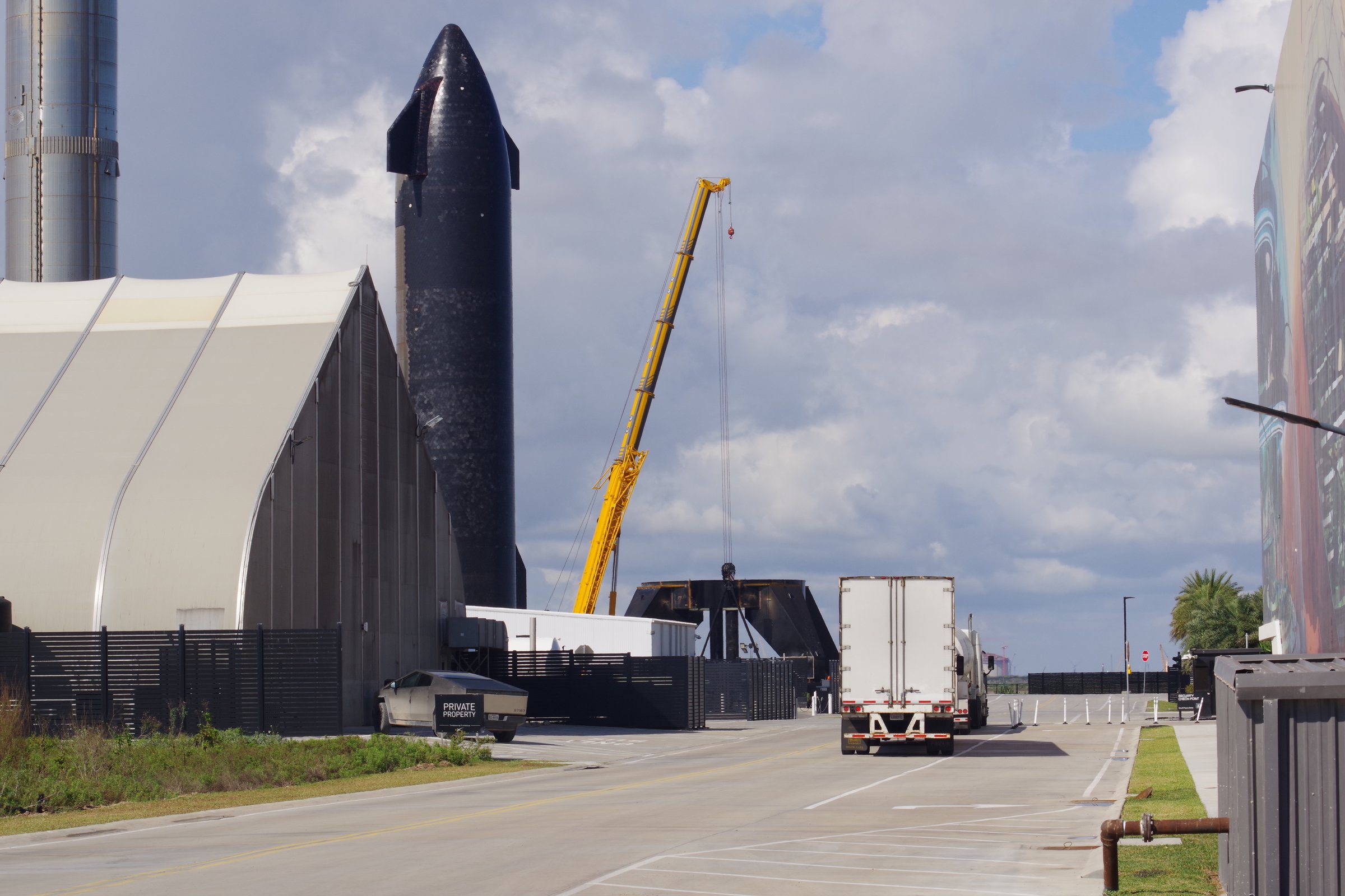 Starship rocket beside a yellow crane and white hangar, Cybertruck by the fence, semi on the road