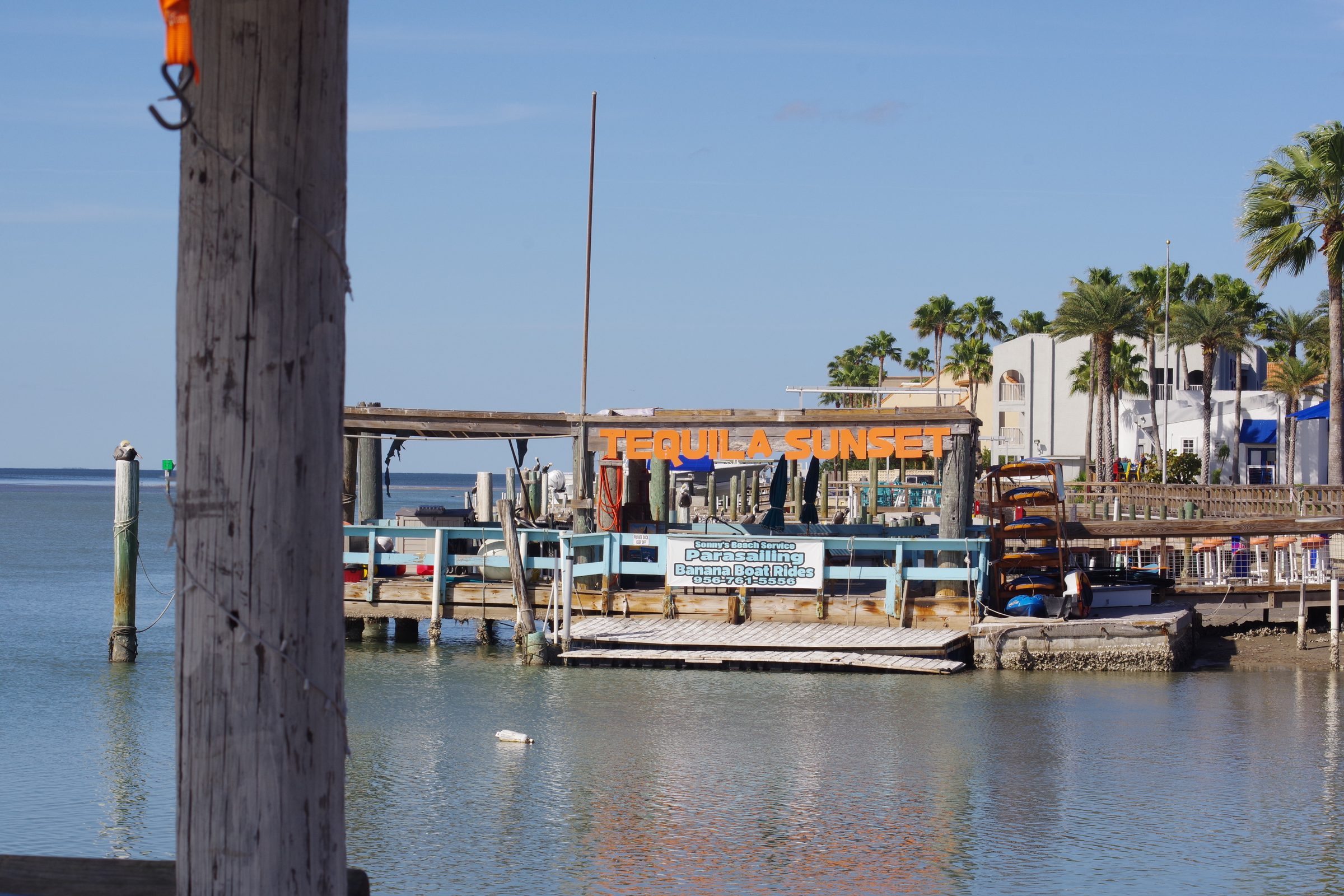 Tequila Sunset dock across the water with kayaks stacked on the pier, palm trees and hotel behind, weathered piling framing left