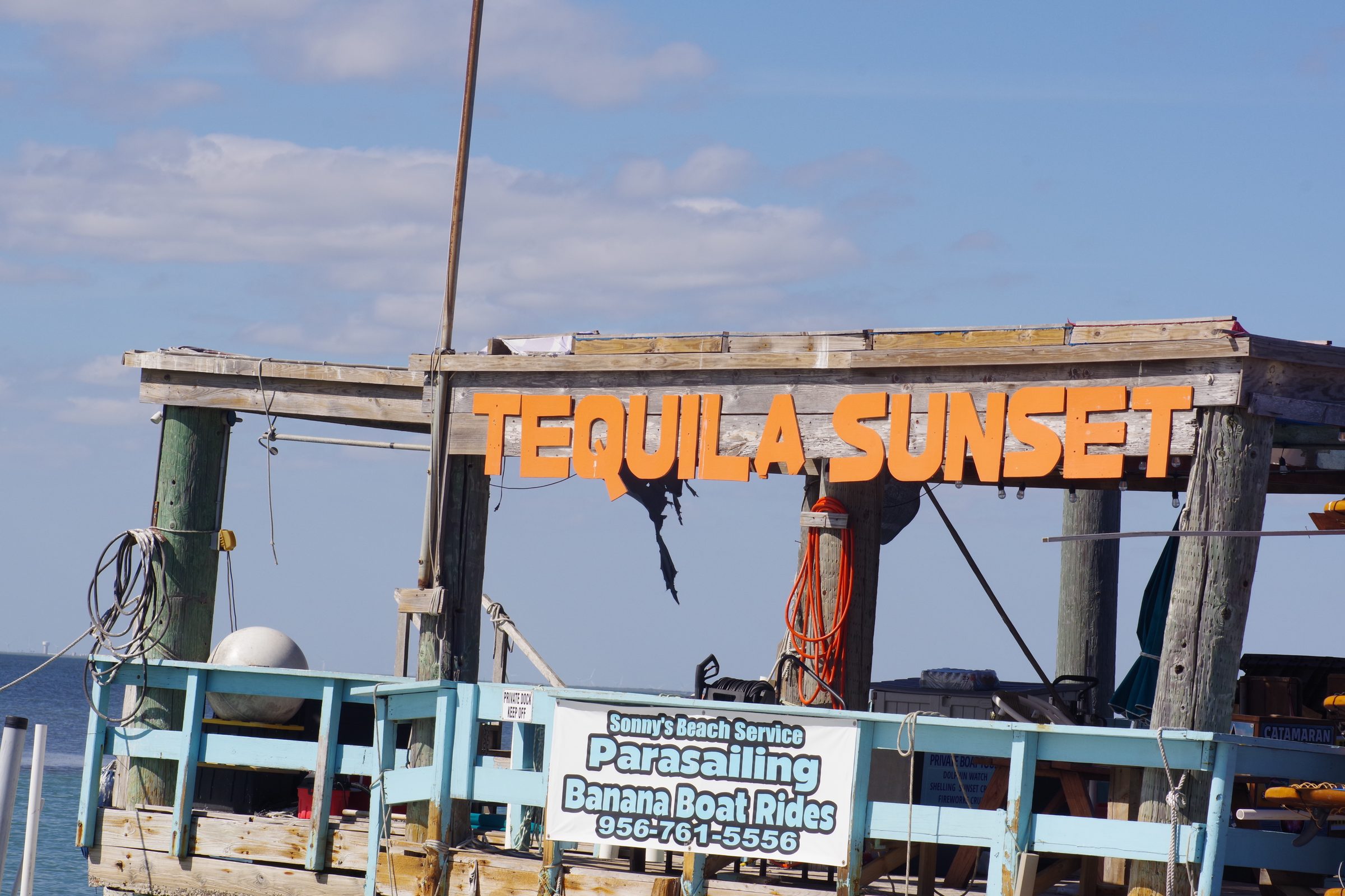 Close shot of weathered dock with large orange TEQUILA SUNSET letters, Sonny's Beach Service banner, orange rope, and blue railing
